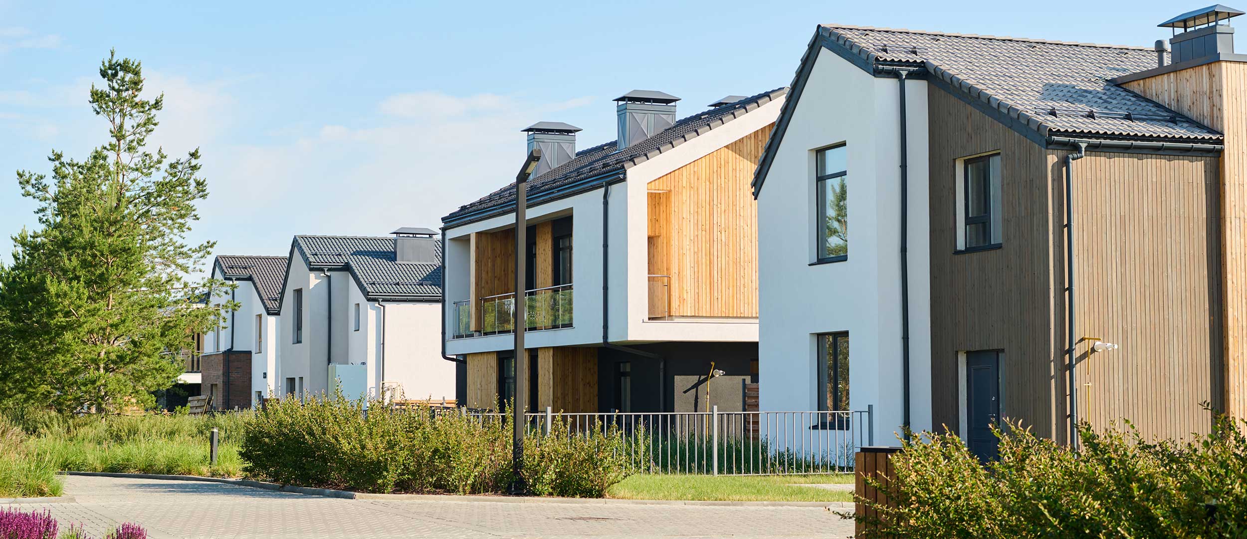 Modern residential buildings with a blend of white and wooden facades, surrounded by greenery and a paved pathway, showcasing design-led property development by Atlas Homes.